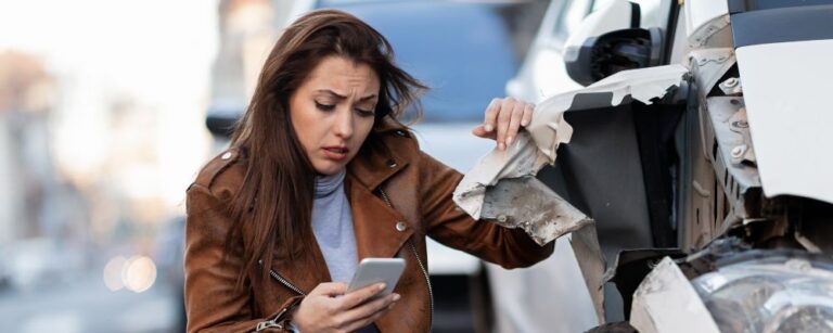 woman with brown hair kneeling next to wrecked car on cell phone
